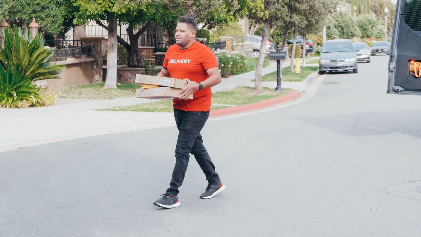 A delivery driver in a Delivery shirt carrying packages down a neighborhood street
